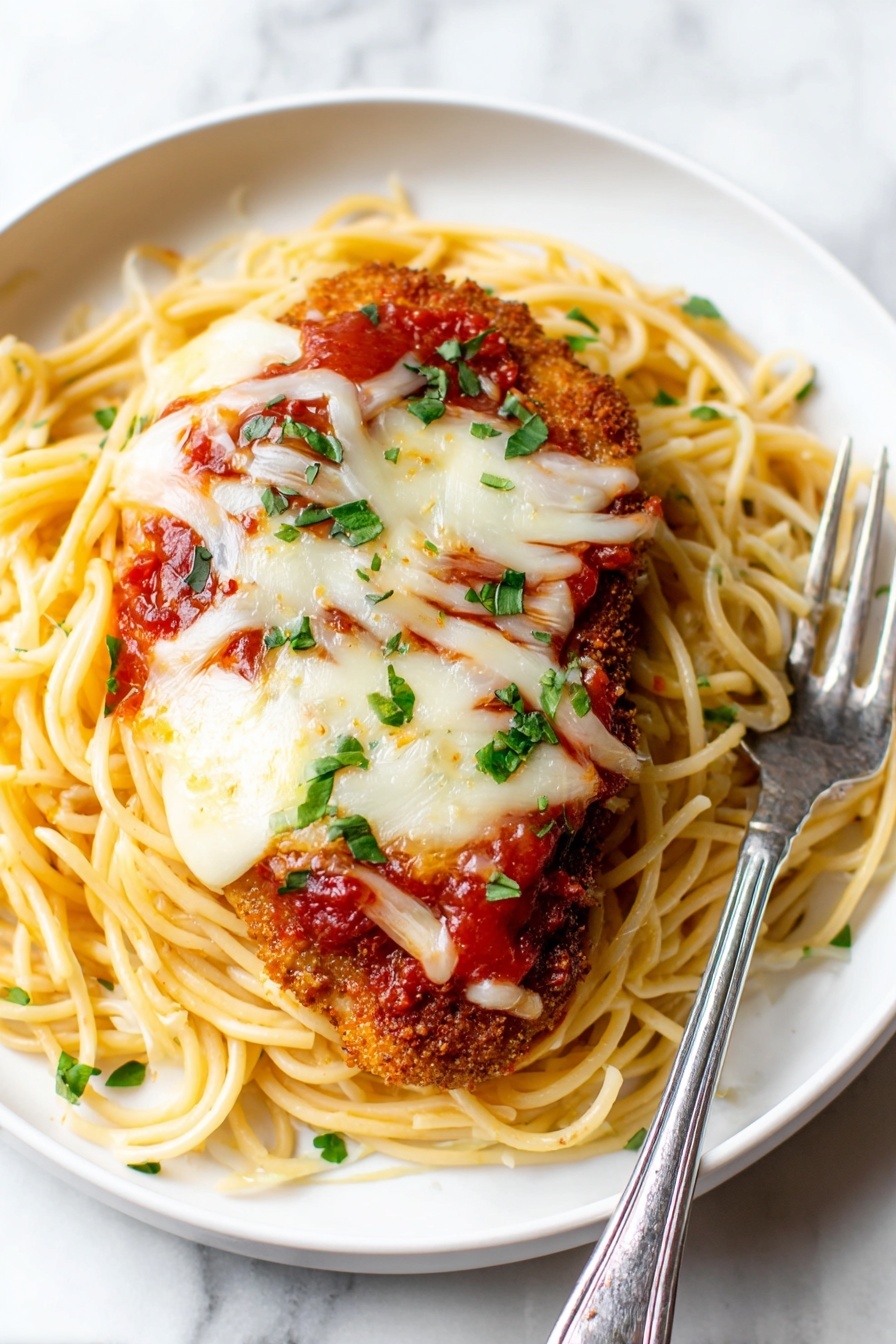 A white plate holds one layer of cooked, slightly shiny spaghetti noodles spread across the base, topped by a second layer of bright red tomato sauce with visible chunks. On top of the sauce sits a breaded, golden-brown piece of chicken covered with a third layer of melted white cheese that looks soft and slightly stretchy. The dish is sprinkled with small bright green chopped herbs for color. A silver fork and spoon rest on the plate’s edge, partially touching the noodles. The plate is set on a white marbled texture. Photo taken with an iphone --ar 2:3 --v 7