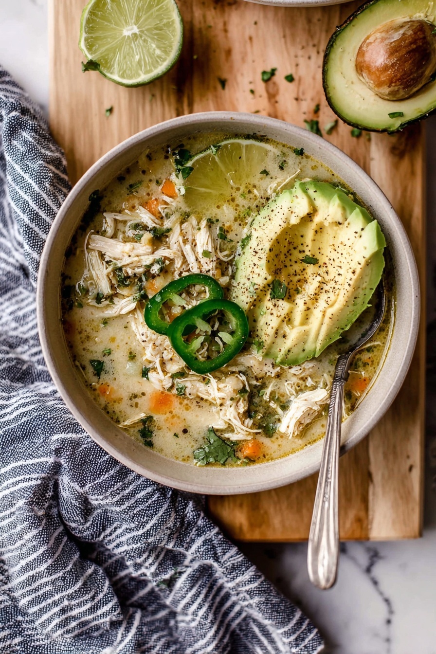 A bowl filled with shredded cooked chicken mixed with a light yellow broth and sprinkled with chopped green herbs and black pepper, layered with bright orange pieces beneath. On top, there are three green jalapeño slices, a fan of pale green avocado slices, and a lime half placed next to them. A silver spoon is placed inside the bowl. The bowl rests on a wooden chopping board, which holds another squeezed lime half. The surface under the board is white marble, and a blue and white striped cloth is partly under the bowl. Fresh green cilantro leaves are scattered around. Photo taken with an iphone --ar 2:3 --v 7