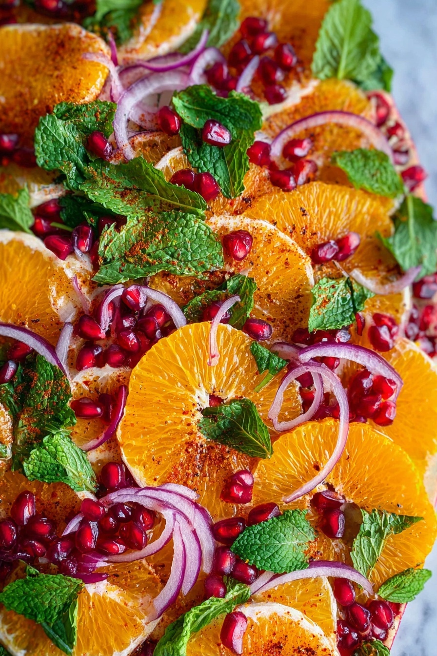 The dish shows many thin orange slices arranged close together as the bottom layer, each slice shiny and slightly juicy with a dusting of red spice on top. Over this, there are scattered thin rings of red onion that are light purple and translucent. Bright green fresh mint leaves are spread evenly across the dish, adding a leafy texture. Small shiny red pomegranate seeds are scattered on top, adding a glossy and round contrast. The entire dish is on a white surface with a white marbled texture. photo taken with an iphone --ar 2:3 --v 7