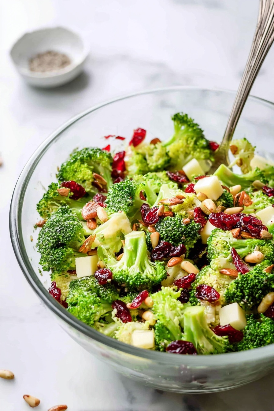 A clear glass bowl is filled with three main layers: bright green broccoli florets with a rough texture form the base, mixed with small cubes of light yellow cheese scattered evenly throughout. On top of the broccoli and cheese, there is a layer of deep red dried cranberries and reddish-brown chopped nuts adding contrast and texture. Sunflower seeds, small and greyish-tan, are sprinkled over the entire salad, adding a subtle crunch. A silver fork is inserted on the right side of the bowl. The bowl rests on a white marbled surface with a small white dish containing some crushed spices in the background, slightly out of focus. Photo taken with an iphone --ar 2:3 --v 7