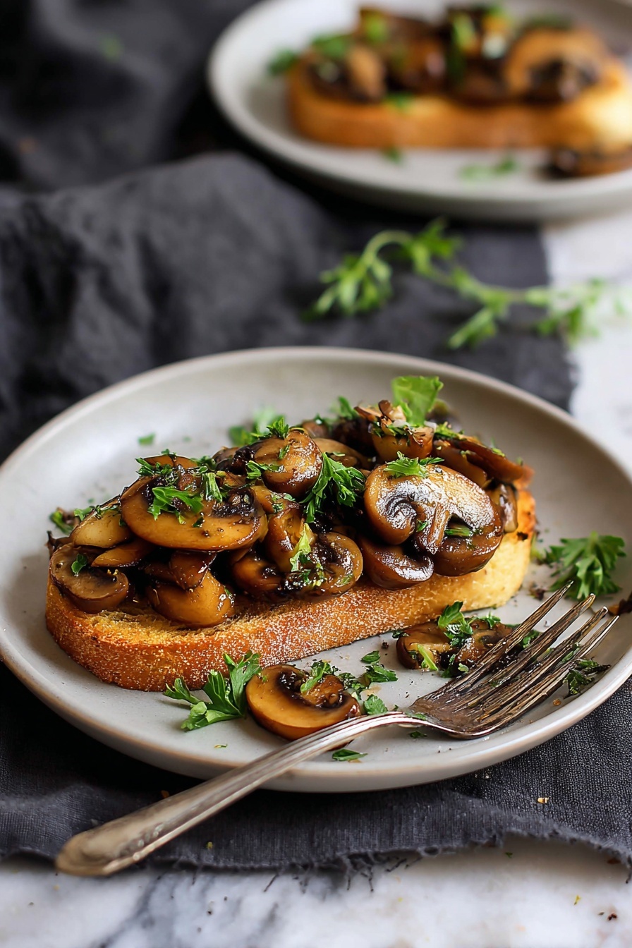 A single slice of golden toasted bread sits in the center of a white plate, topped with a generous layer of sautéed brown mushrooms that have a glossy, slightly crispy texture. Bright green chopped herbs are sprinkled on top and around the plate, adding fresh pops of color. A silver fork rests on the plate's edge, and some mushrooms lie beside the toast. The plate is set on a dark gray cloth on a white marbled surface, with a blurred second plate with a similar dish in the background. photo taken with an iphone --ar 2:3 --v 7