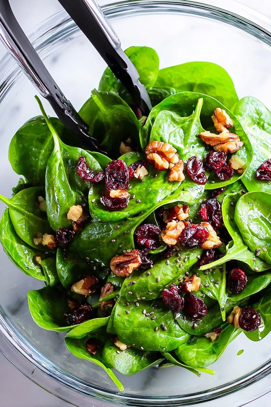 The image shows a clear glass bowl filled with fresh green spinach leaves as the base layer. Scattered evenly across the spinach are small dark red dried cranberries and light brown chopped walnuts, creating small clusters of texture and color. Tiny black chia seeds are sprinkled throughout the salad, adding a dotted texture on top of the spinach and toppings. Stainless steel salad tongs with black silicone tips rest inside the bowl, positioned at the top left side. The bowl sits on a white marbled surface, highlighting the vibrant colors of the salad. photo taken with an iphone --ar 2:3 --v 7