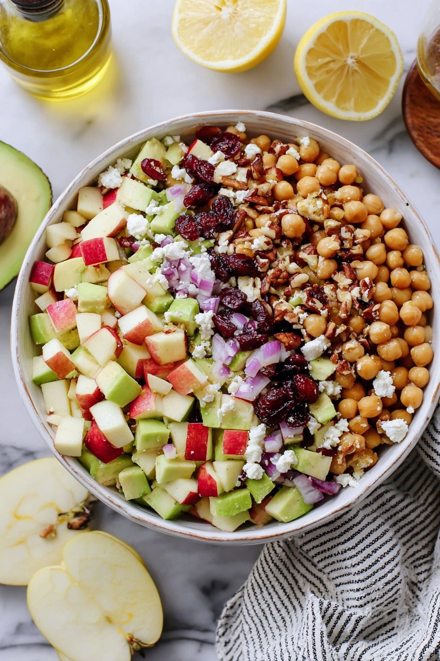A bowl filled with a colorful salad sits on a white marbled surface. The salad has several layers: at the bottom are light beige chickpeas, mixed with chunks of red and green apple pieces that have both smooth and slightly rough textures. Scattered throughout are bright green avocado chunks with a creamy texture, purple-red onion cubes adding a sharp contrast, and small dark red dried cranberries giving a chewy look. On top there are crumbled white feta cheese pieces and some small brown pecans adding a crunchy texture. Three lemon wedges with pale yellow flesh rest on the side of the bowl. A striped white and black cloth is partially visible near the bowl, along with half of a lemon and some slices of green apple in the background. Photo taken with an iphone --ar 2:3 --v 7