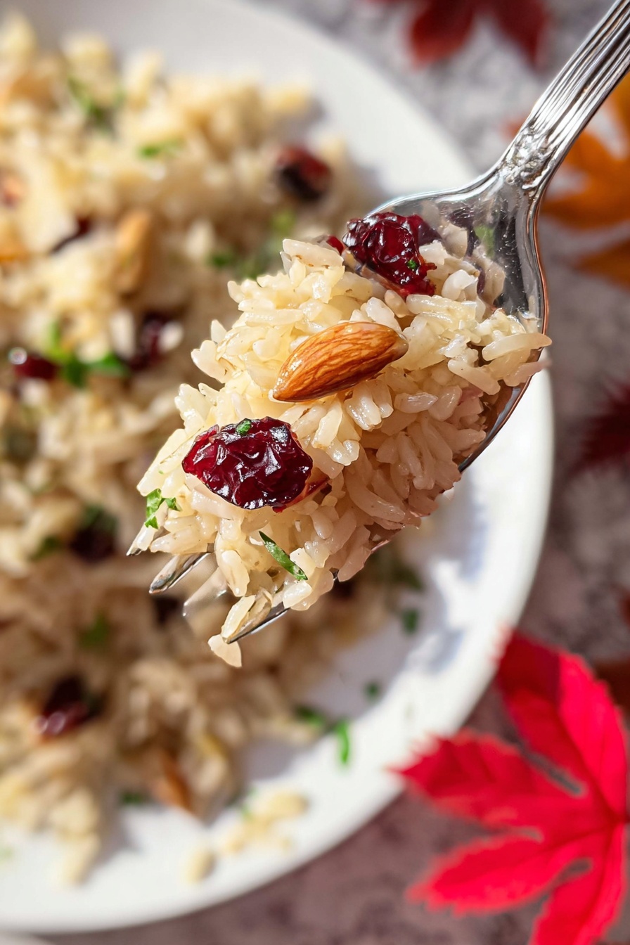 The image shows a close-up of a fork holding a small mound of light brown rice with visible textures of soft, cooked grains. Mixed in the rice are dark red dried cranberries and a small piece of beige almond, adding color contrast. In the blurred background, a white plate holds a larger serving of the same rice mix, garnished with a few small green herb leaves. The scene rests on a white marbled surface with scattered artificial red autumn leaves adding a warm tone to the setting. Photo taken with an iphone --ar 2:3 --v 7