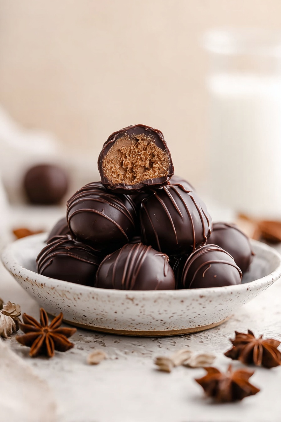 A small white speckled plate holds nine round chocolate truffles stacked in a pile, with one truffle at the top cut in half to show a smooth light brown filling inside. Each truffle is coated with dark chocolate and has thin lines of darker chocolate drizzled on top, creating textured patterns. The plate is placed on a white marbled surface, with a soft warm beige background blurred behind it. Star anise pieces are scattered around the plate, and a glass of milk stands slightly blurred in the back left. photo taken with an iphone --ar 2:3 --v 7