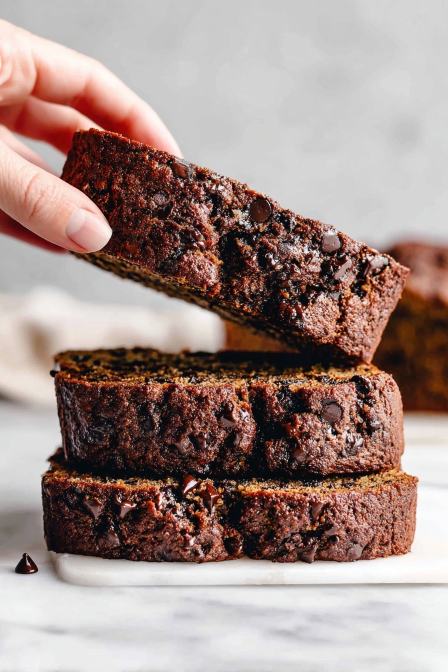 There are three slices of dark brown chocolate chip bread stacked on a white marbled surface, with the top slice being lifted by a woman's hand. The bread looks moist with many small, dark chocolate chips scattered throughout each slice, showing a rich texture. In the background, more slices are slightly blurred. Photo taken with an iphone --ar 2:3 --v 7