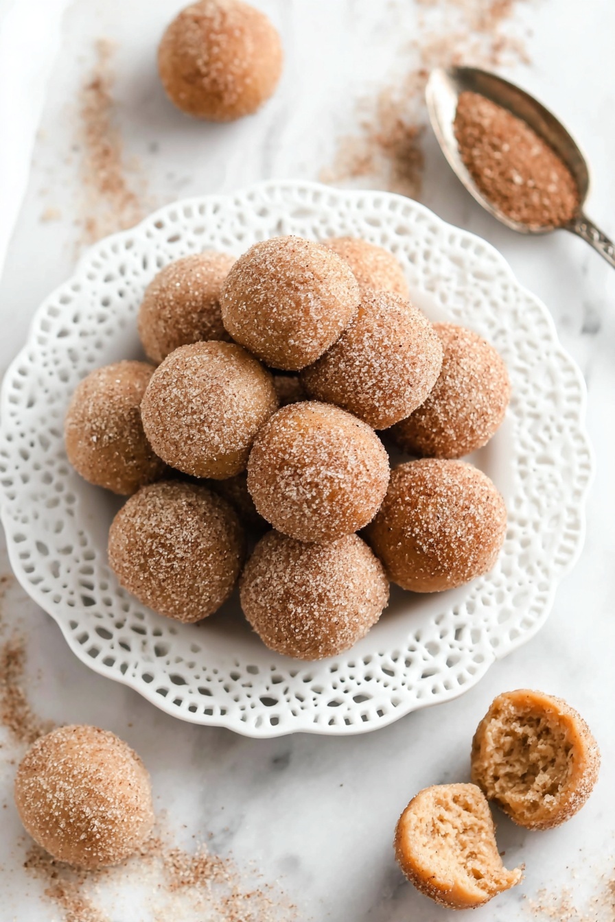 A white plate with lace edges is piled high with round, sugar-coated mini donuts. The donuts have a light golden brown color with a textured surface, dusted evenly with cinnamon sugar. One donut is broken open at the front, showing a soft, fluffy inside with a pale beige color. Around the plate, there are a few more mini donuts scattered on a white marbled surface, along with a silver spoon holding some of the cinnamon sugar mix. In the background, there is a blur of a glass cup with a light brown drink. photo taken with an iphone --ar 2:3 --v 7