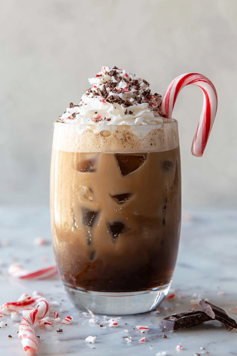 The image shows a clear glass filled with three visible layers: the bottom layer is dark chocolate brown, the middle layer is a lighter brown, and the top layer is a creamy mix with ice cubes inside. On top, there is a swirl of white whipped cream with small chocolate flakes sprinkled over it. A red and white striped candy cane is hooked on the rim of the glass. Around the glass, on a white marbled surface, there are small pieces of broken chocolate and crushed candy cane scattered. Photo taken with an iphone --ar 2:3 --v 7