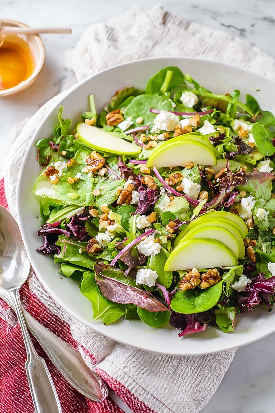 This image shows a white plate filled with a fresh salad layered with bright green apple slices on top, mixed vibrant green spinach and arugula leaves as the base layer, and scattered pieces of soft white cheese crumbles. There are also chunks of reddish-brown dried fruit and small pieces of red onion visible throughout the salad. Light brown walnut pieces are sprinkled partly hiding under the leaves, adding texture. A silver fork and spoon rest on the left side of the plate. The plate sits on a white marbled surface with a blurred wooden board and green apple halves in the background. Photo taken with an iphone --ar 2:3 --v 7