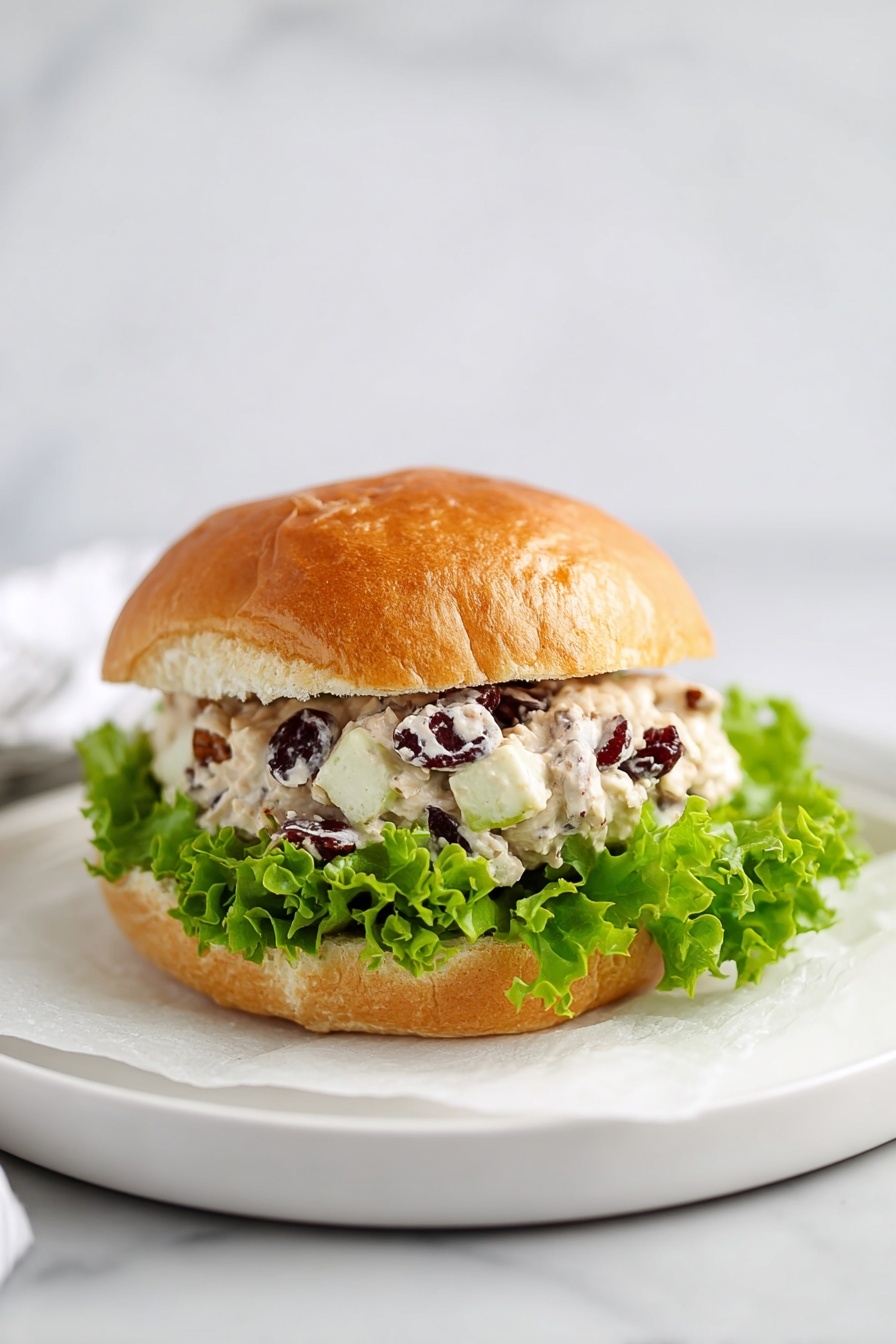 A sandwich on a soft and golden-brown round bun with a slightly cracked top. The bottom layer holds fresh, bright green lettuce leaves with curly edges. Above the lettuce is a creamy salad filling made of white chunks, possibly chicken or cheese, mixed with small brown nut pieces and some reddish bits, all coated in a light, creamy dressing. The top half of the bun rests lightly on the filling. The sandwich sits on a white plate lined with white paper, placed on a white marbled surface. photo taken with an iphone --ar 2:3 --v 7