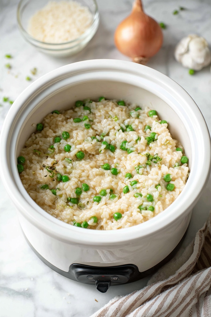 A white bowl filled with creamy risotto that is mixed with bright green peas, showing a soft and slightly shiny texture. The risotto has small bits of onion and is topped with a light sprinkling of black pepper. Behind the bowl is a clear glass bowl filled with uncooked rice. To the left, there is a whole garlic bulb on a white marbled surface. Some pieces of shaved cheese are scattered on the surface near the bowl, with a block of cheese partly visible on the corner. To the right side, a white rice cooker with