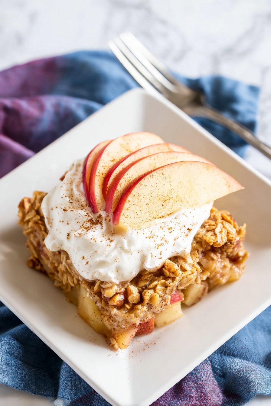 The dish features a single thick layer of baked oatmeal with visible chunks of apples and walnuts, creating a mix of light yellow, red, and brown textures. On top of this base layer, three thin slices of red apple are arranged in a slight fan shape. A generous dollop of white whipped cream sits atop the apple slices, sprinkled with a light dusting of cinnamon powder. The oatmeal is served on a white square plate placed on a white marbled surface, with a fork and two folded cloth napkins in muted purple and blue colors nearby. photo taken with an iphone --ar 2:3 --v 7