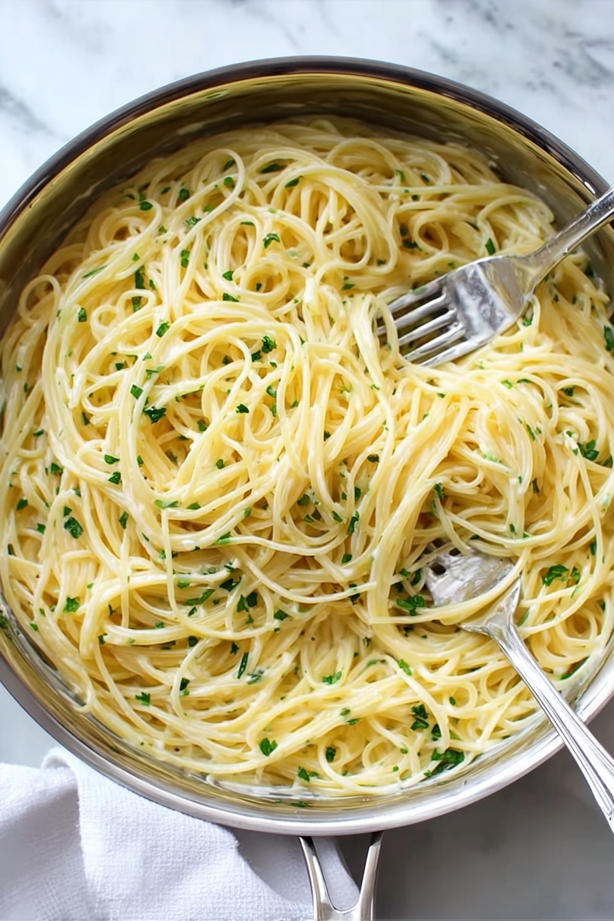 A close-up top view of a silver pan filled with creamy spaghetti pasta mixed with finely chopped green herbs scattered evenly throughout. Two silver forks are twirling strands of the light yellow pasta in the center, showing the smooth and shiny texture of the sauce coating each strand. The pan sits on a soft white cloth on a white marbled surface, giving a clean and fresh look. Photo taken with an iphone --ar 2:3 --v 7