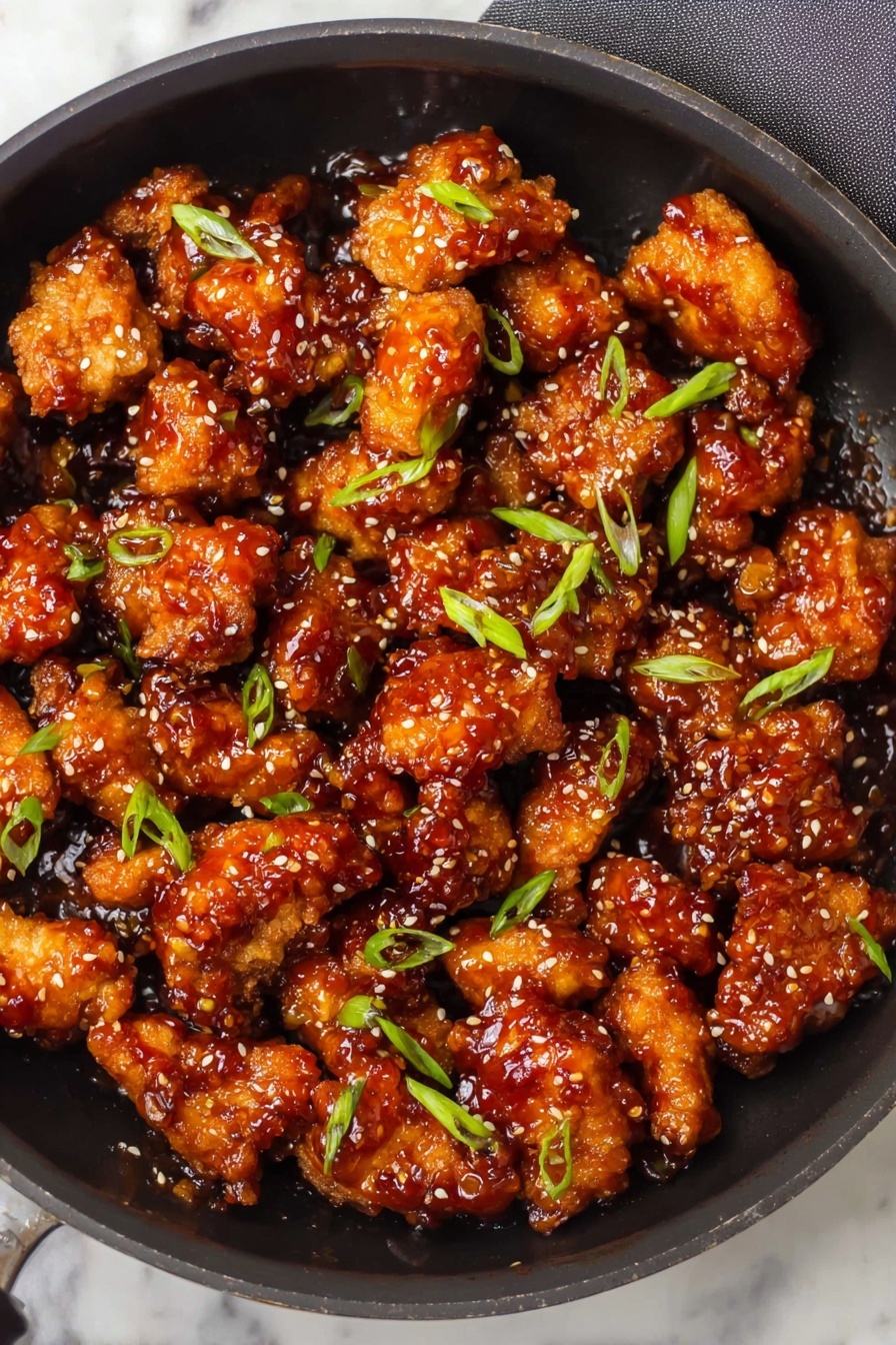 A black pan filled with many pieces of golden brown chicken coated in a shiny reddish-brown sauce, sprinkled evenly with small white sesame seeds and small green sliced scallions. The chicken pieces vary in size and texture, looking crispy with a sticky glaze, and some edges appear slightly darker from cooking. The pan handle is visible on the right side, and the background is a white marbled texture. photo taken with an iphone --ar 2:3 --v 7