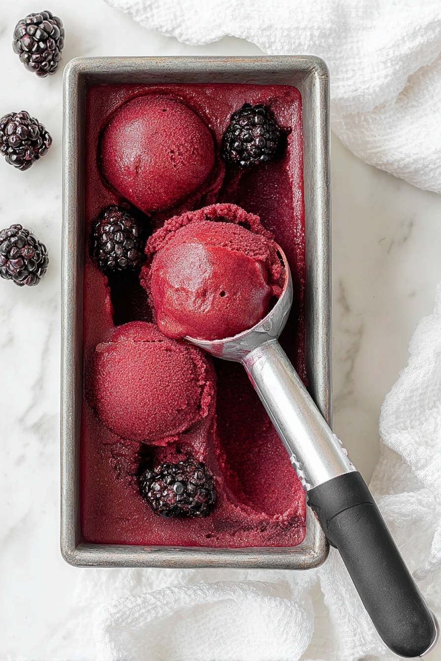 The image shows a rectangular metal container filled with deep red blackberry sorbet, smoothly textured with some uneven scooped sections. Three scoops of sorbet sit on top, two resting in the middle and one held by a black-handled scoop near the bottom left. Around the edges and in the sorbet are whole blackberries, dark purple-black in color and glossy. The container sits on a white marbled surface with a white cloth underneath, and more blackberries are scattered on the surface. Photo taken with an iphone --ar 2:3 --v 7